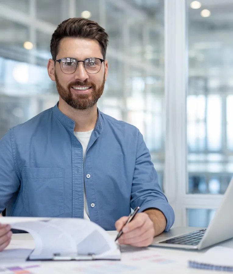 A smiling businessman in a blue shirt and glasses sits at a desk, reviewing documents with a pen in hand, alongside a laptop in a bright modern office.