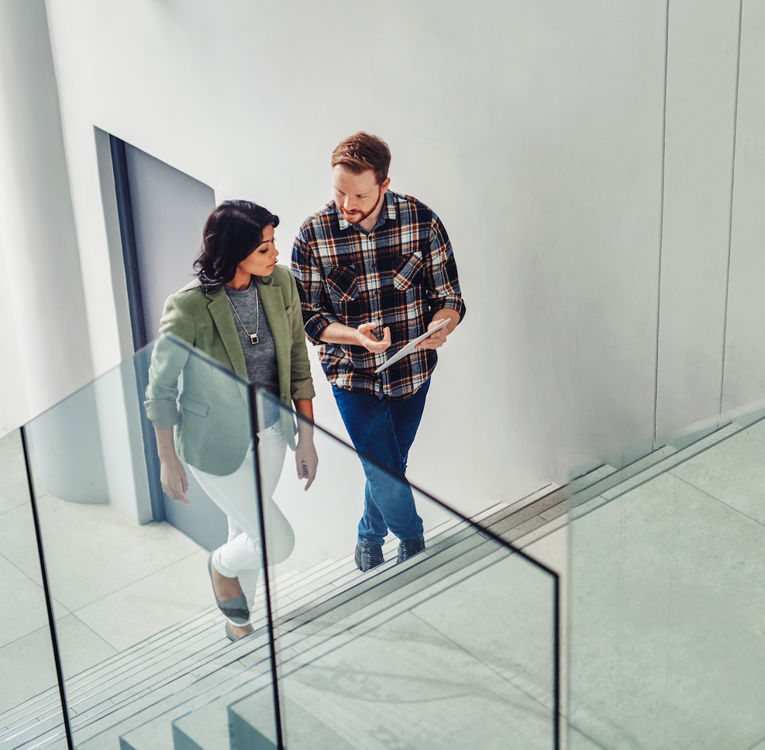 A man in a plaid shirt holding a tablet and a woman in a green blazer are engaged in a conversation while walking up a modern staircase with glass railings. The setting suggests a collaborative and dynamic work environment. Ideal for themes like teamwork, workplace communication, and informal business meetings