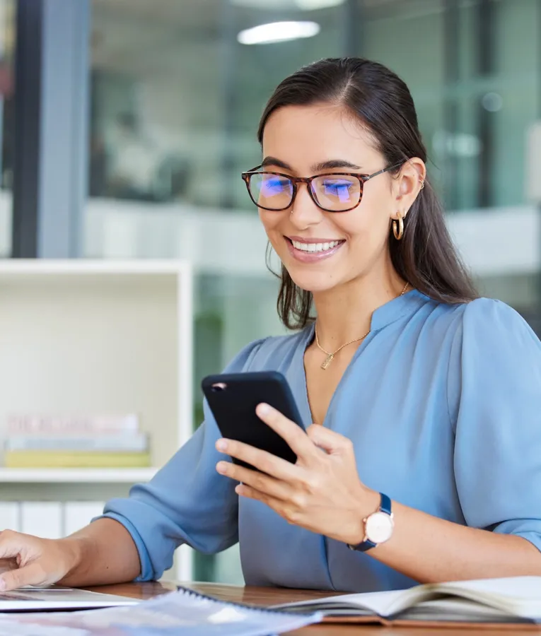 Smiling office professional in blue blouse using smartphone and laptop at desk, representing digital workforce communication, hybrid work, and productivity in a modern workplace.