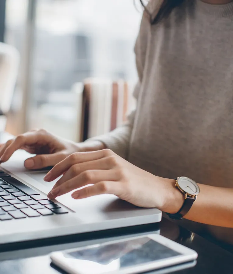 A remote worker in a beige sweater types on a laptop at a modern workspace. A smartphone lies on the dark wooden table, reflecting a flexible and digital work environment.