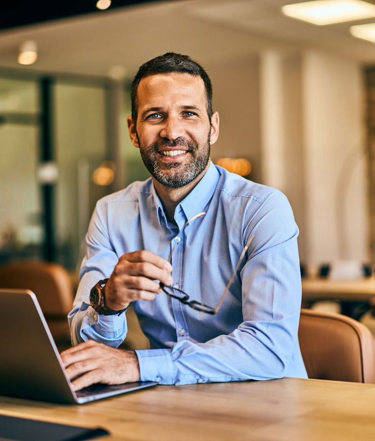 Smiling businessman holding a cup of coffee while working on a laptop in a modern office setting.