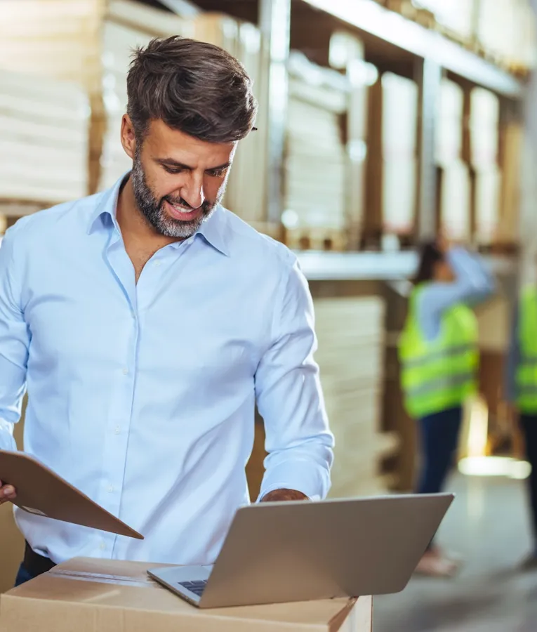 A warehouse manager in a light blue shirt holding a clipboard and checking inventory on a laptop, with workers in high-visibility vests in the background.