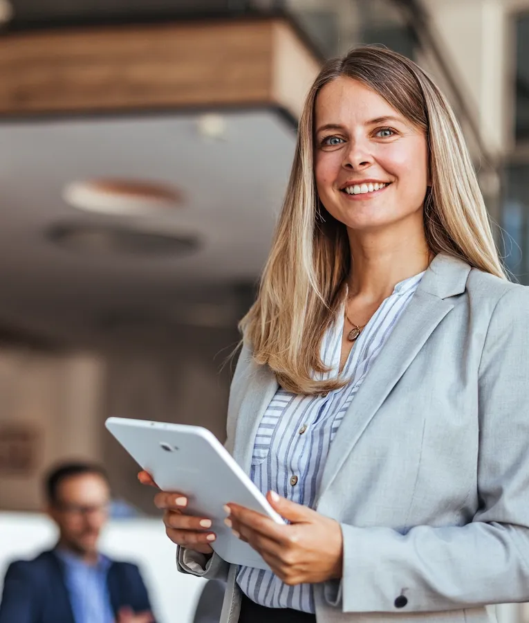 A smiling businesswoman in a light grey blazer and striped blouse holds a tablet while standing in a modern office with colleagues in the background.