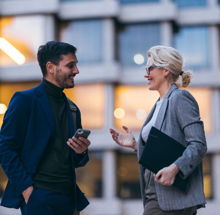 A man in a blue suit holding a smartphone and a woman in a gray blazer holding a folder are having a friendly conversation outside an office building. The background features illuminated windows, creating a modern business atmosphere. Ideal for themes like networking, collaboration, and workplace communication.