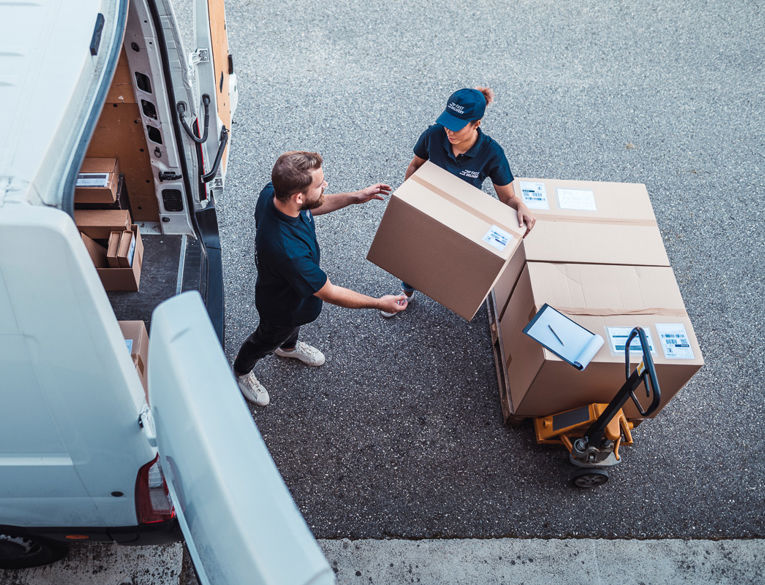 Two delivery workers in navy uniforms unloading large cardboard boxes from a van onto a pallet with a clipboard nearby.