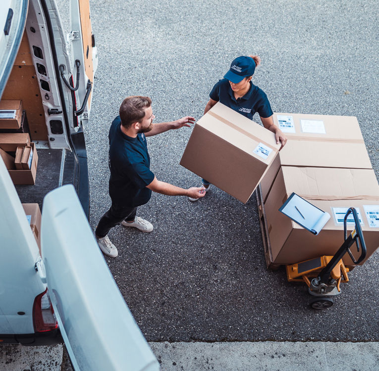 Two delivery workers in navy uniforms unloading large cardboard boxes from a van onto a pallet with a clipboard nearby.