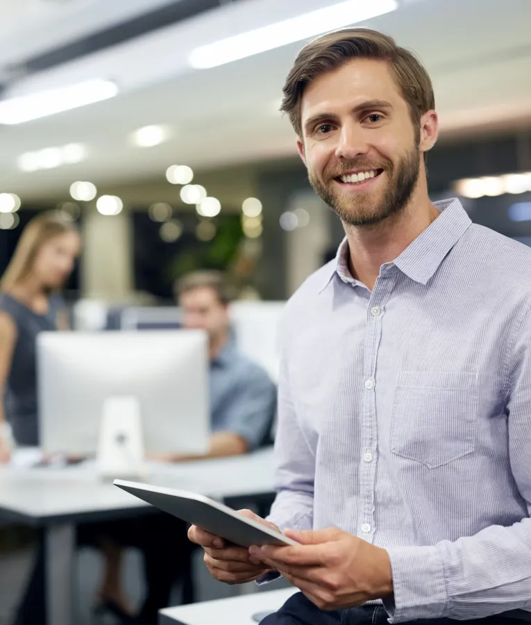 Smiling businessman in a modern office holding a tablet, with colleagues working in the background. Represents efficient workforce management and digital collaboration.