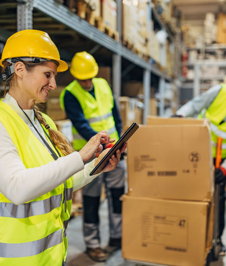 A smiling female warehouse worker in a high-visibility vest and hard hat using a tablet to check inventory, while colleagues organize boxes in the background.