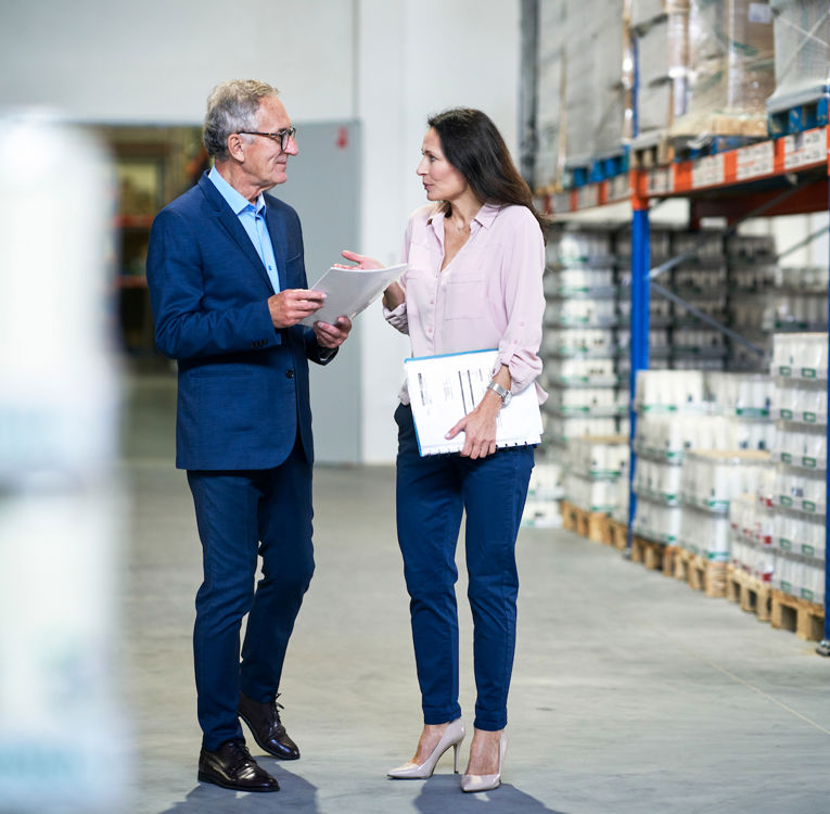 Two business professionals discussing logistics in a warehouse while holding documents and a tablet.