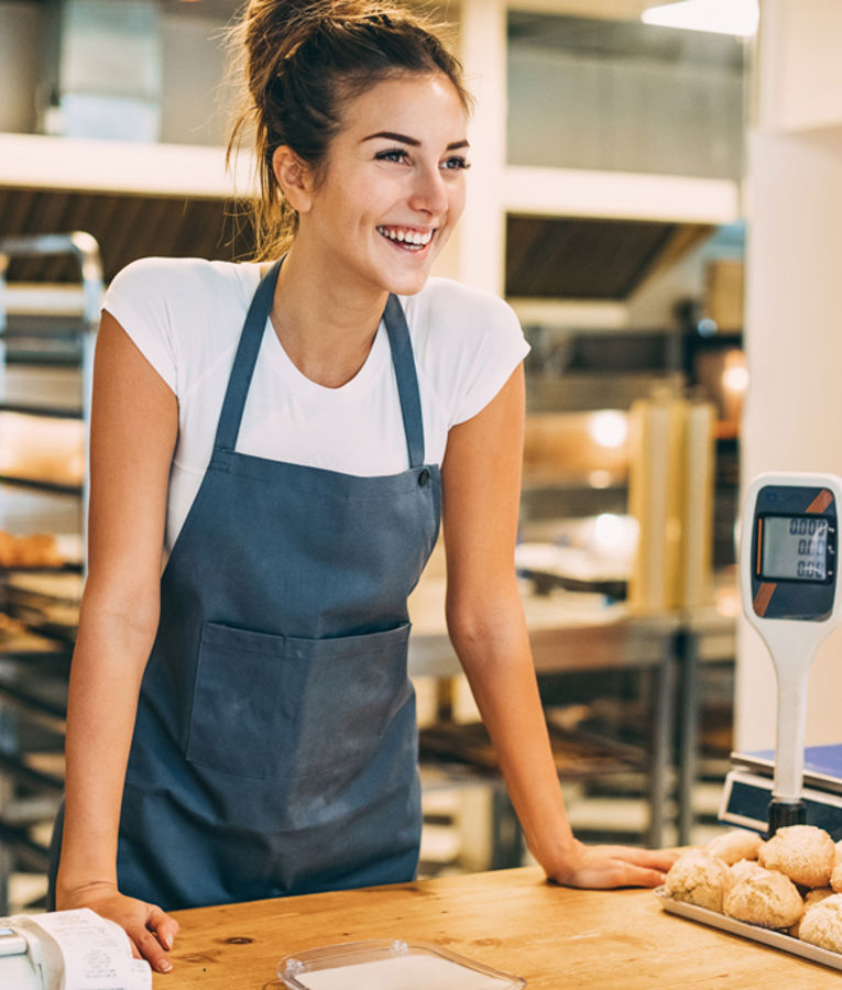 Smiling bakery employee wearing apron standing behind the counter with bread rolls and a digital scale, representing retail service, shift scheduling, and customer engagement.