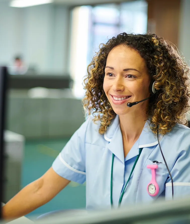 A smiling nurse in a light blue uniform with a headset assisting patients at a hospital reception desk.