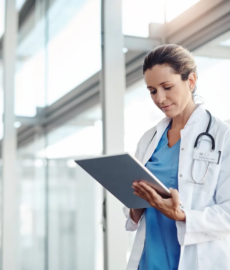 A female doctor in a white lab coat with a stethoscope carefully reviewing patient data on a tablet in a bright, modern hospital corridor.