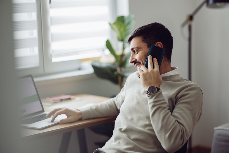 Man working from home with a laptop and phone, managing staff scheduling and time tracking through an AI-based workforce management solution.