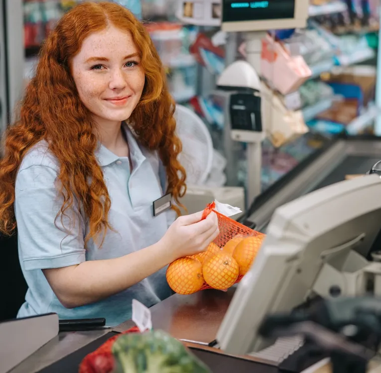 Smiling young supermarket cashier scanning oranges at checkout, representing retail workforce efficiency, POS operation, and customer service.