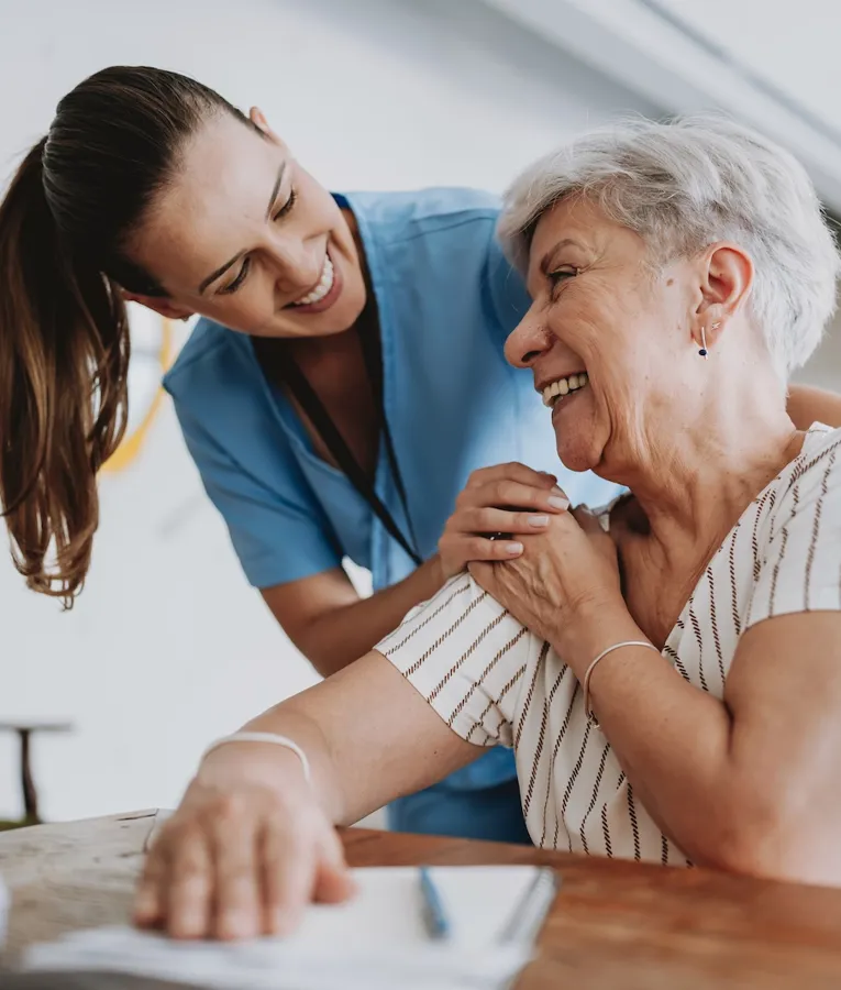 A smiling caregiver in blue scrubs warmly embracing a happy elderly woman at a care facility.