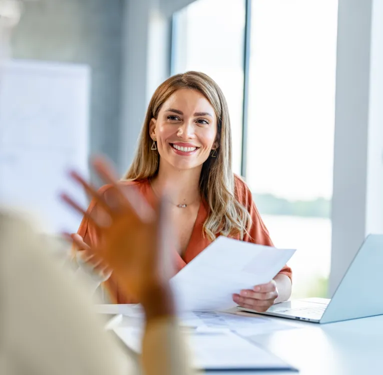 A smiling workforce manager in a burnt-orange blouse holds documents while engaging in a discussion with an employee in a bright, modern office setting.