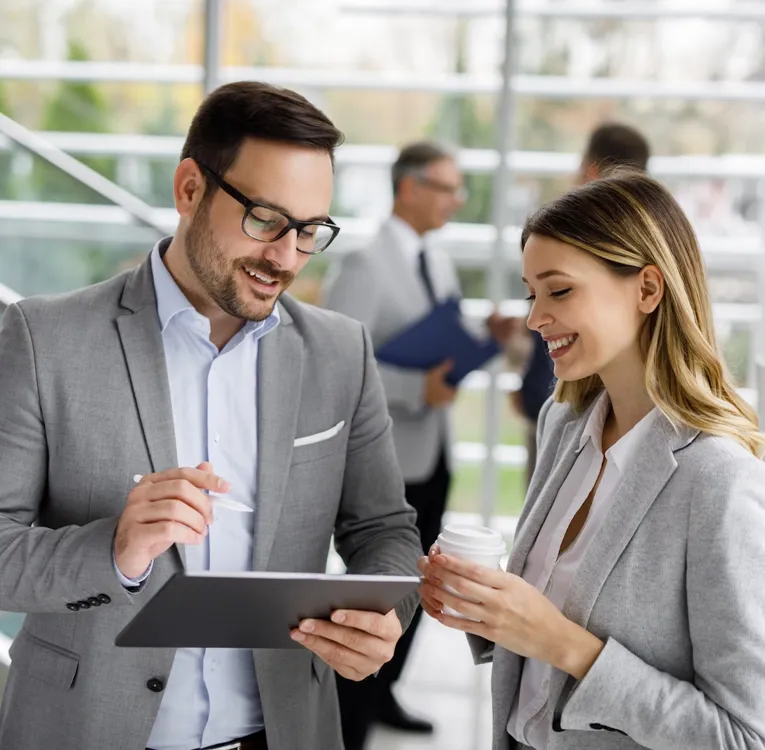 A smiling businessman and businesswoman in grey suits discuss workforce strategies while reviewing data on a tablet in a modern office setting.