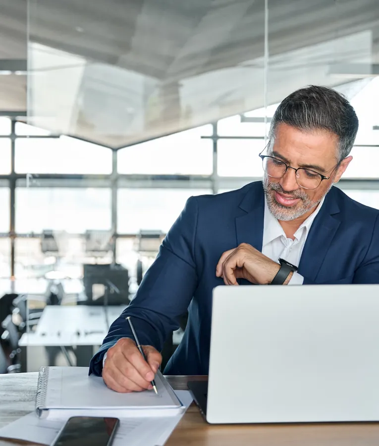A professional middle-aged man in a navy blue suit working on a laptop in a modern office. He is taking notes in a notebook while checking his smartwatch, suggesting efficient time management and productivity.
