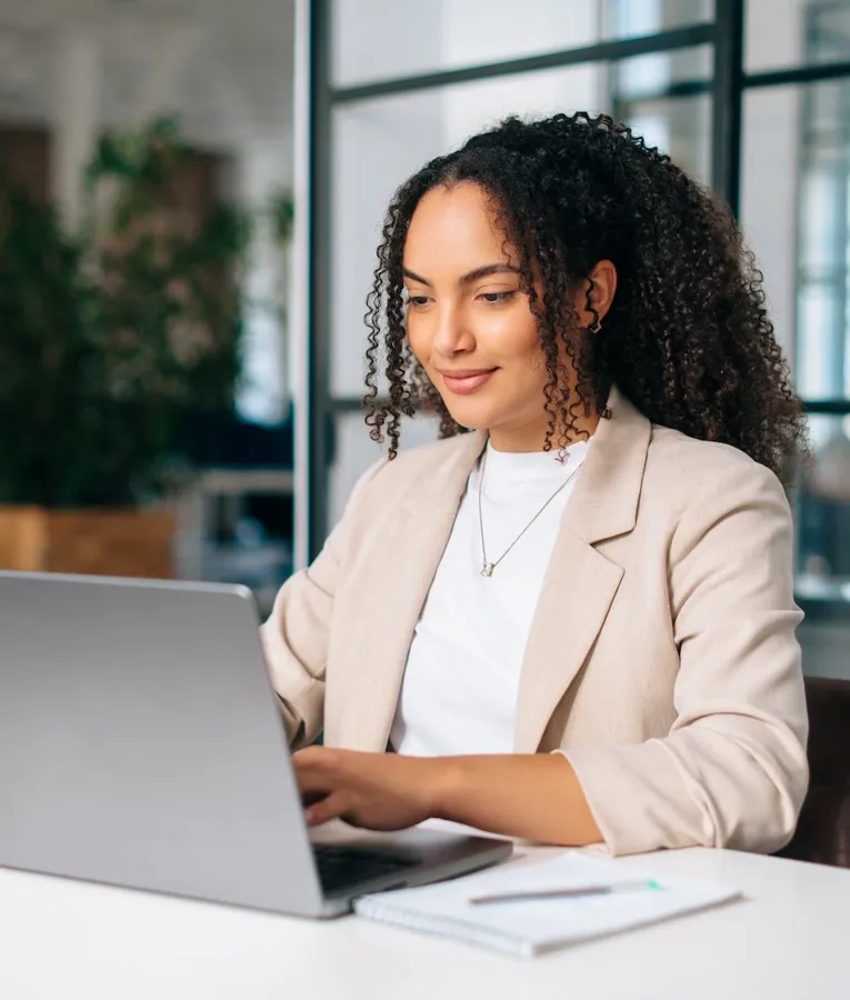 A young professional in a beige blazer works on a laptop at a bright and modern office desk, surrounded by notes and office supplies, representing productivity and focus.