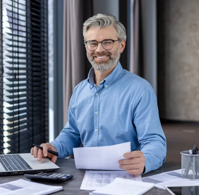 Smiling business professional in blue shirt working with documents and laptop at office desk, representing data analysis, workforce reporting, and financial planning.