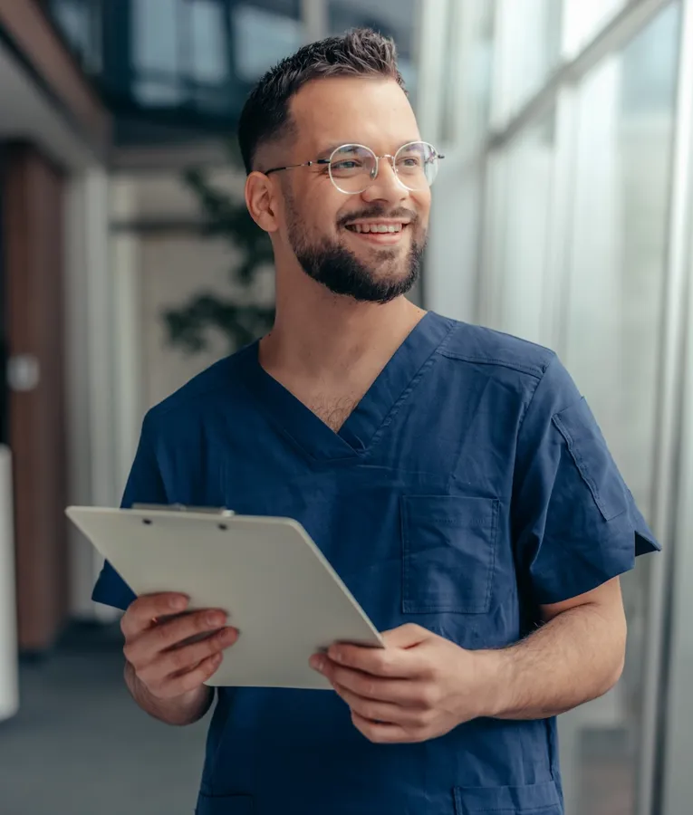 A smiling male nurse in navy blue scrubs holding a clipboard while standing in a bright hospital corridor.