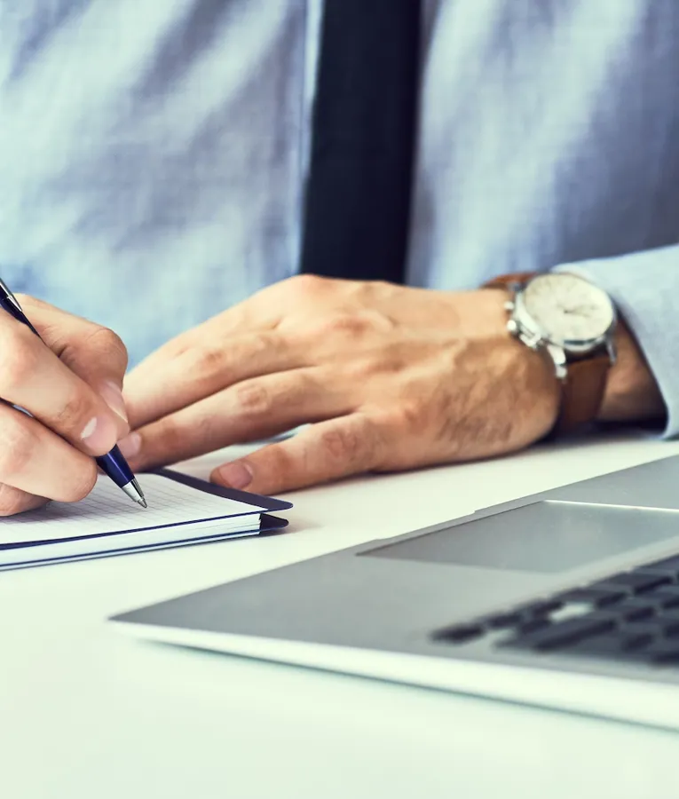 A close-up of a businessman in a blue shirt and tie writing notes in a notebook while working on a laptop at a modern office desk.