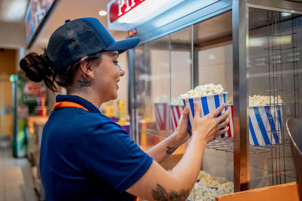Smiling cinema employee in uniform placing a striped popcorn bucket into a display warmer, representing shift work, retail service roles, and frontline workforce efficiency.