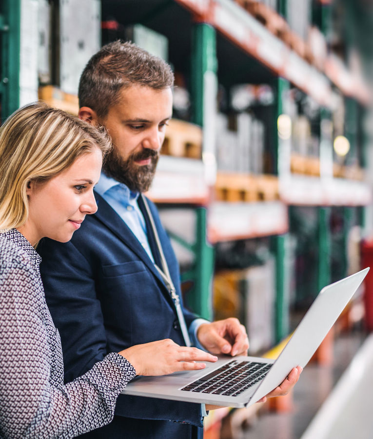 Two professionals collaborating on a laptop in a warehouse, discussing inventory management strategies.