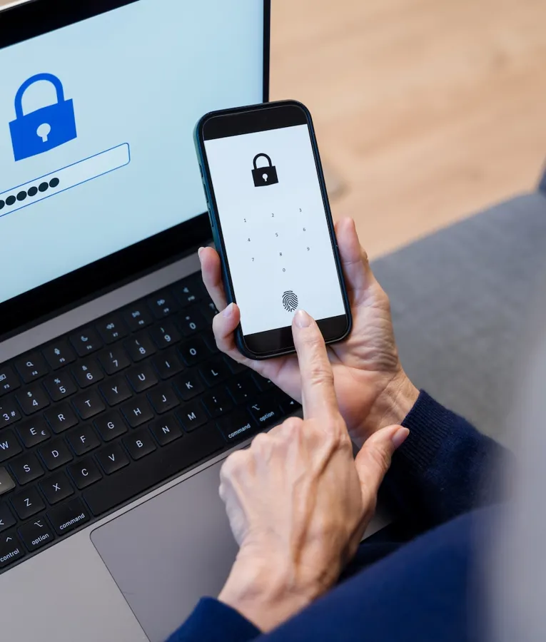 A person using biometric authentication on a smartphone while logging into a laptop with a password screen displaying a security lock icon.