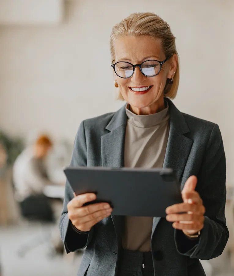 A smiling senior businesswoman in a grey suit and glasses holds a tablet while working in a modern office. Colleagues are collaborating in the background, reflecting a productive and dynamic workplace.