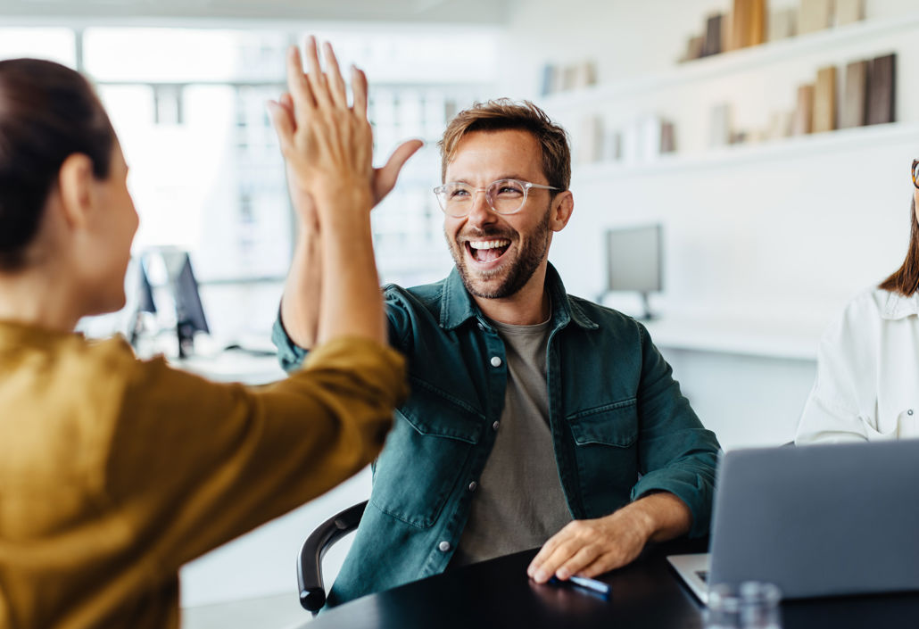 Two colleagues celebrating with a high five in the office, symbolising strong team spirit, employee engagement, and satisfaction with collaborative scheduling tools.