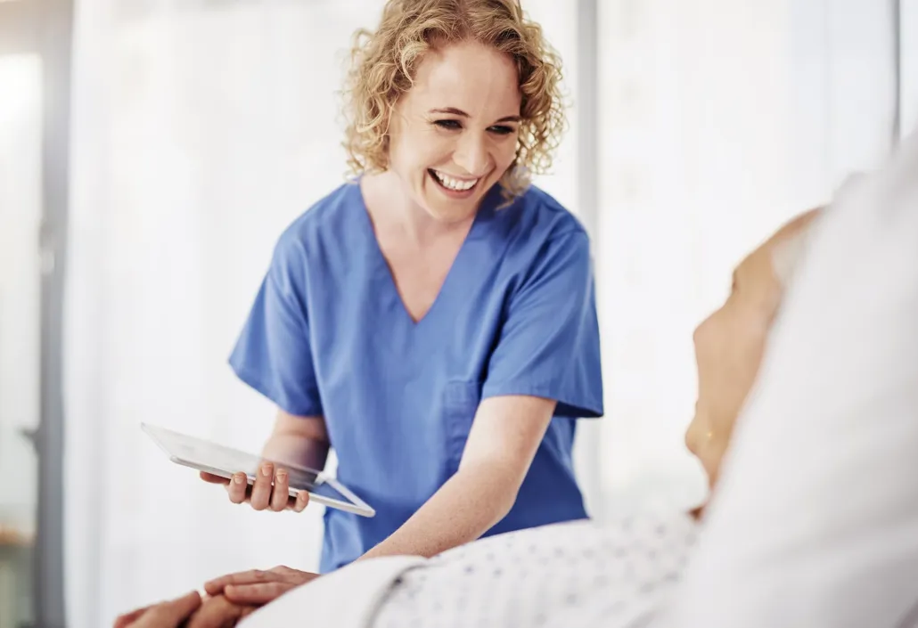 A smiling healthcare worker uses a tablet at a patient’s bedside – representing efficient time tracking and shift planning in the care sector.