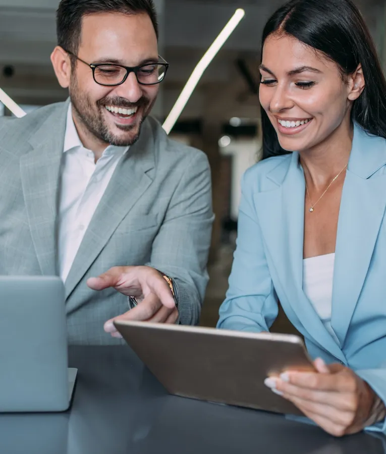 A smiling businesswoman in a light blue blazer holds a tablet while collaborating with a male colleague in a grey suit, who is pointing at a laptop screen. They are in a modern office setting, discussing workforce management strategies.