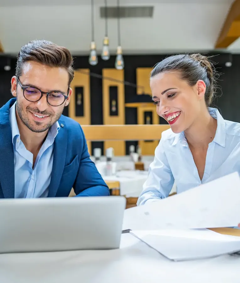 A smiling man in a blue suit and a woman in a light blue blouse collaborate on a laptop and review documents in a modern restaurant setting.