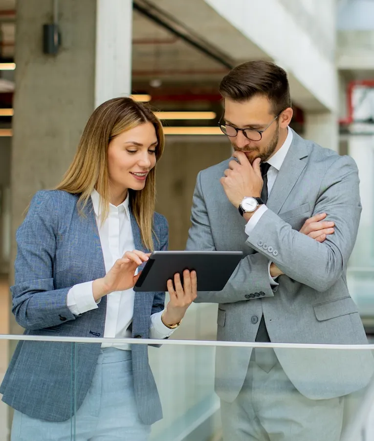 A businesswoman in a blue blazer presenting information on a tablet to a thoughtful male colleague in a modern office hallway.