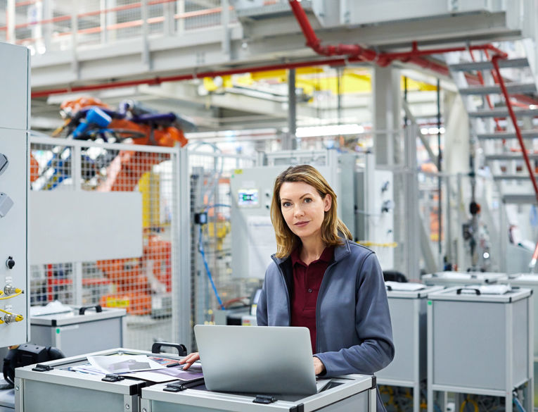 A confident female engineer working on a laptop in a modern industrial facility, monitoring automated production systems.