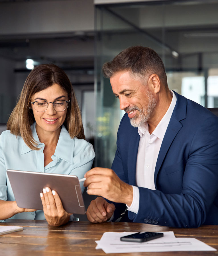 Two business professionals collaborating in a modern office, reviewing information on a tablet. The woman wears glasses and a light blue shirt, while the man, dressed in a navy suit, smiles and gestures toward the screen. Ideal for workforce management, teamwork, or corporate strategy content.