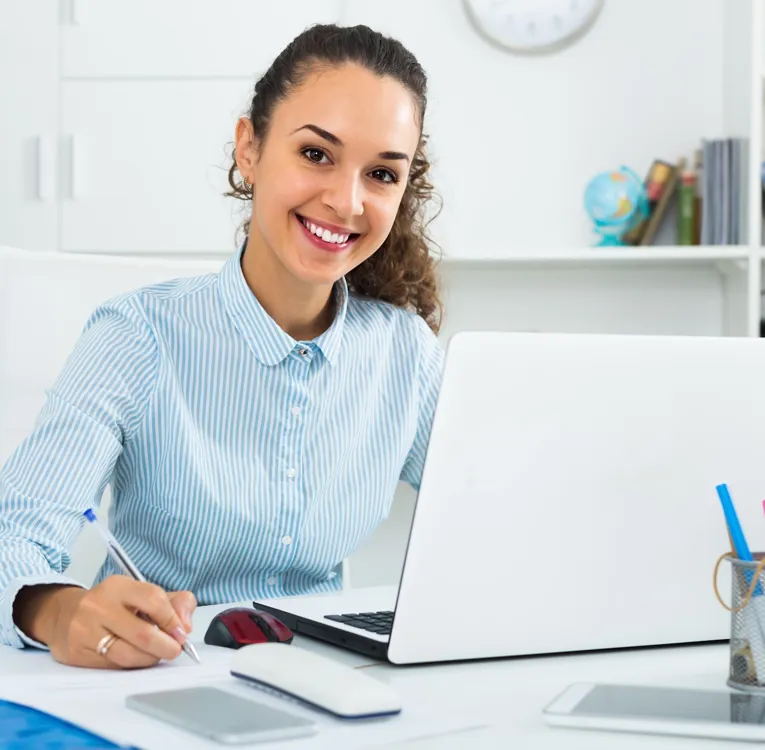 Happy office worker in striped shirt writing notes at desk with laptop, showcasing workforce coordination, task planning, and digital administration.