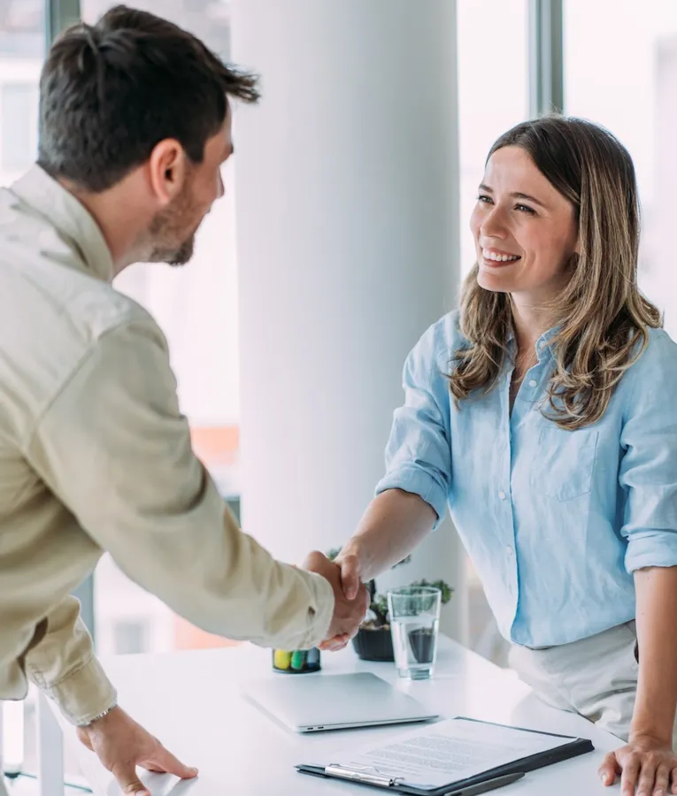 Smiling business professionals shaking hands across a desk in a bright office, symbolising successful collaboration, onboarding, and positive workplace culture.