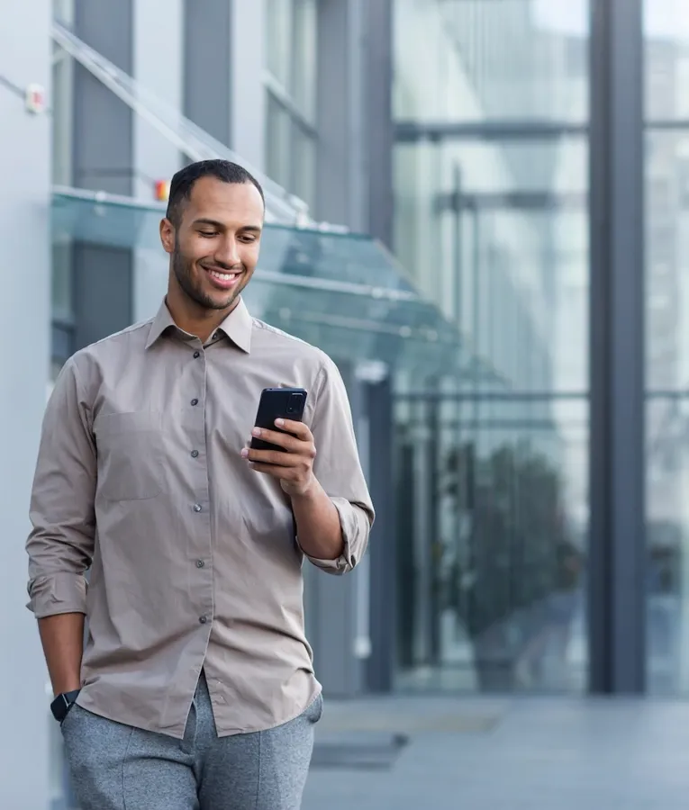 A smiling professional in a beige shirt walks outside a modern glass office building while checking his smartphone, representing a mobile and flexible work environment.