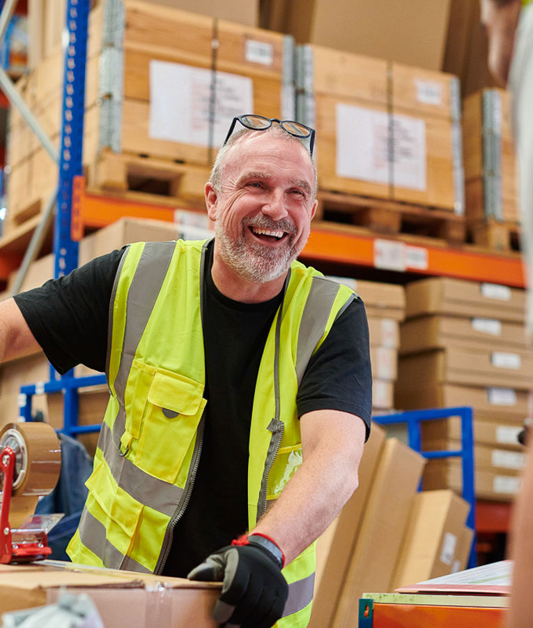 A smiling warehouse worker in a high-visibility vest and gloves packing boxes in a busy storage facility.